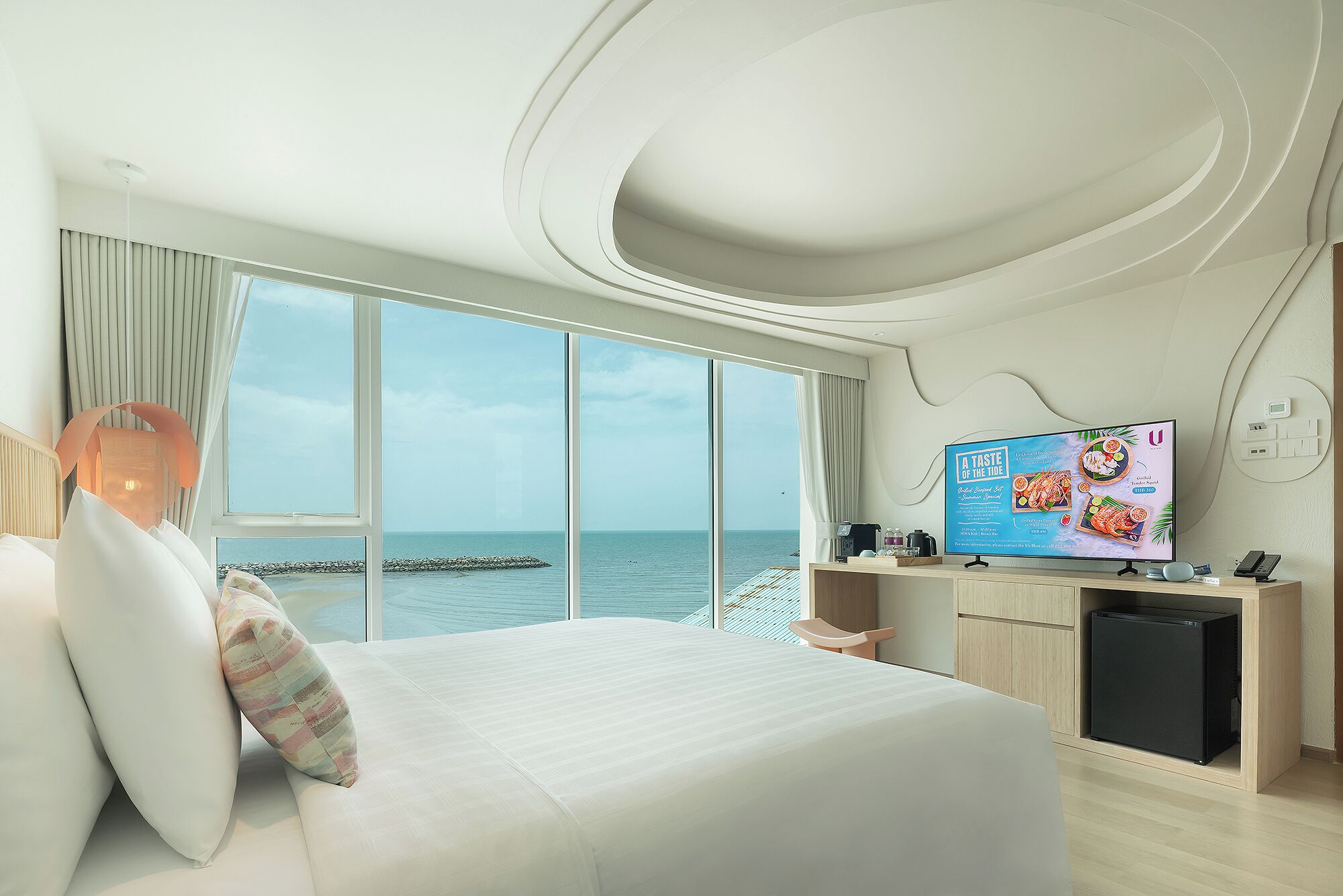A modern hotel room with a large bed, a TV displaying a menu, and a view of the sea through large windows.
