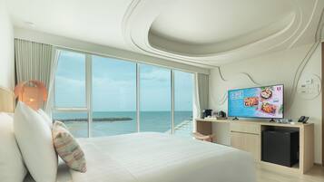 A modern hotel room with a large bed, a TV displaying a menu, and a view of the sea through large windows.