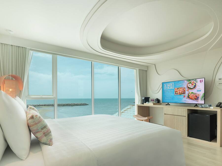 A modern hotel room with a large bed, a TV displaying a menu, and a view of the sea through large windows.
