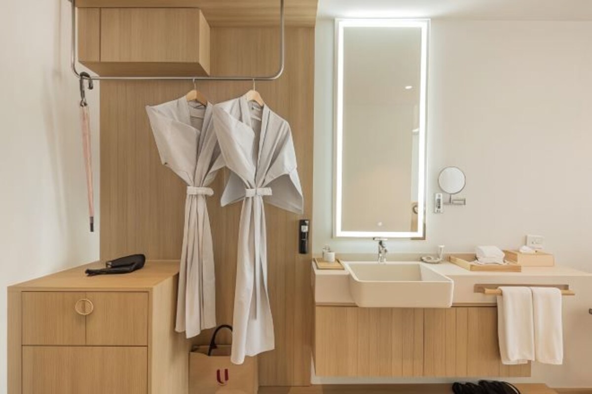 A modern bathroom with wooden cabinetry, a large mirror, and two white robes hanging on a rack.