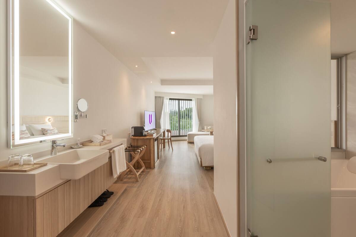 Une chambre d’hôtel moderne dotée d’un grand miroir, d’une coiffeuse en bois, d’un bureau avec une chaise, d’un lit et d’une cabine de douche en verre.