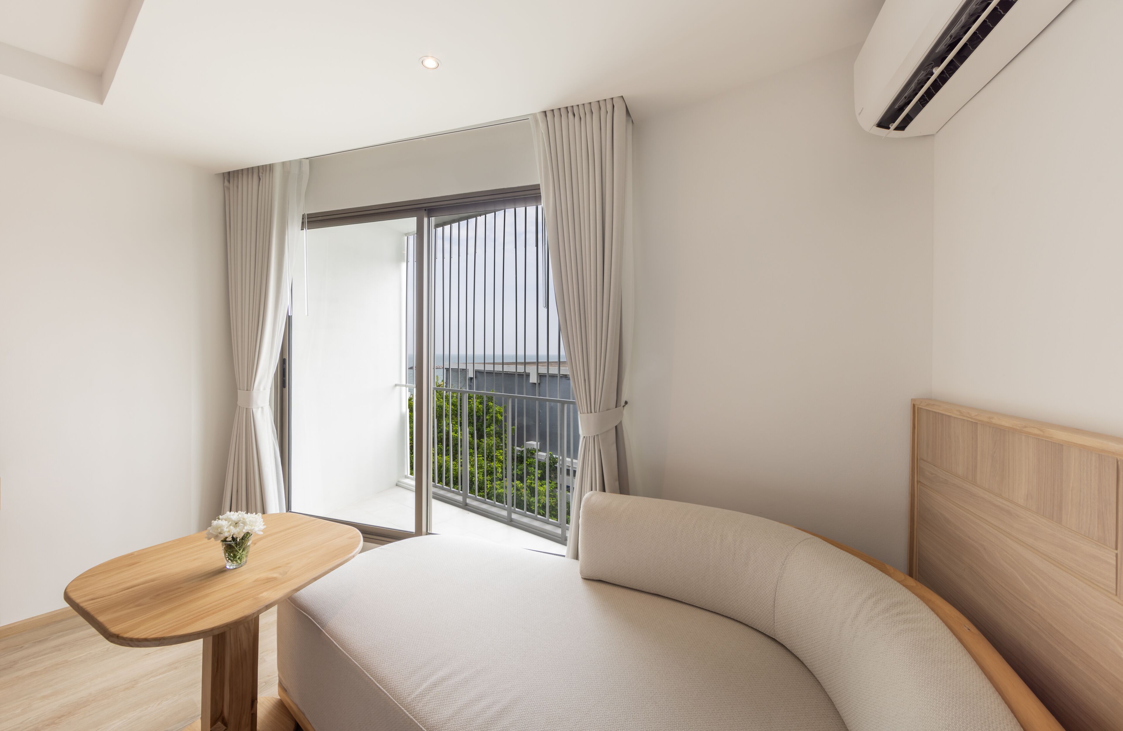 Een moderne slaapkamer met een gebogen bed, een klein houten tafeltje en een groot raam met gordijnen.