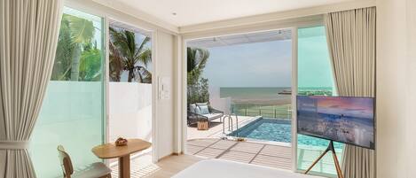 Habitación de hotel con una cama grande, televisión, zona de piscina con vista a la playa y comedor con mesa y sillas.