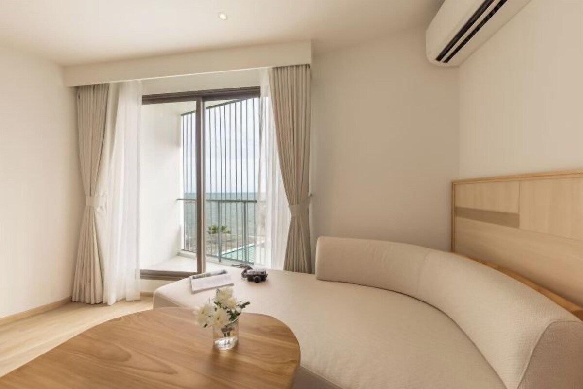 A modern hotel room with a curved sofa, a wooden coffee table with a vase, and a view of a balcony.