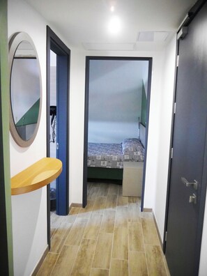 Un estrecho pasillo que conduce a un dormitorio con una cama, un pequeño estante de madera y una puerta.