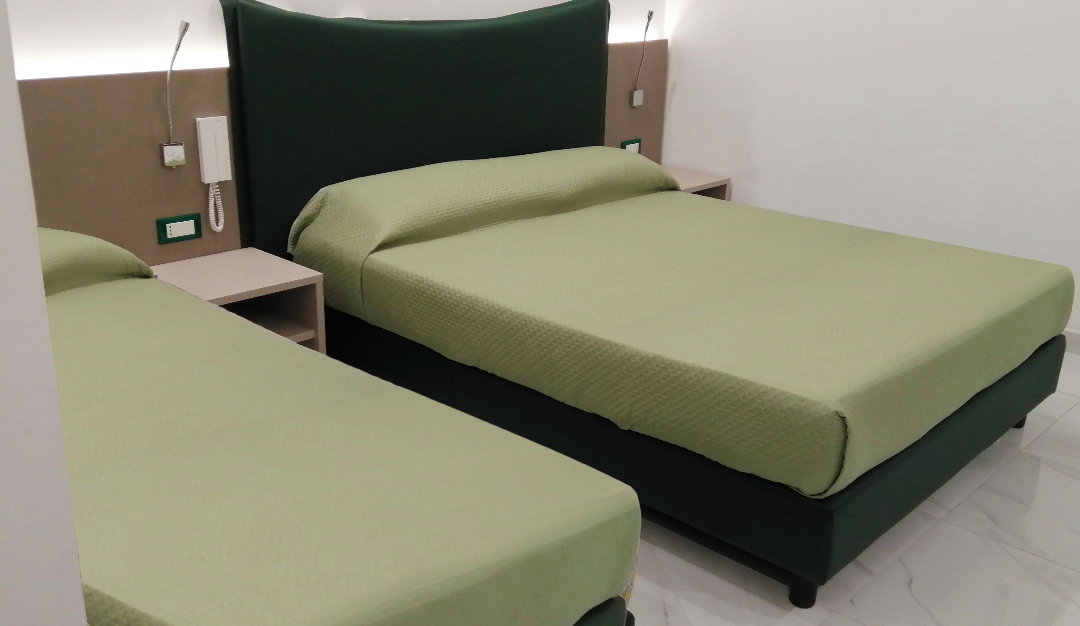 A hotel room with two single beds, each with a green bedspread, a wooden nightstand, and a wall-mounted telephone.