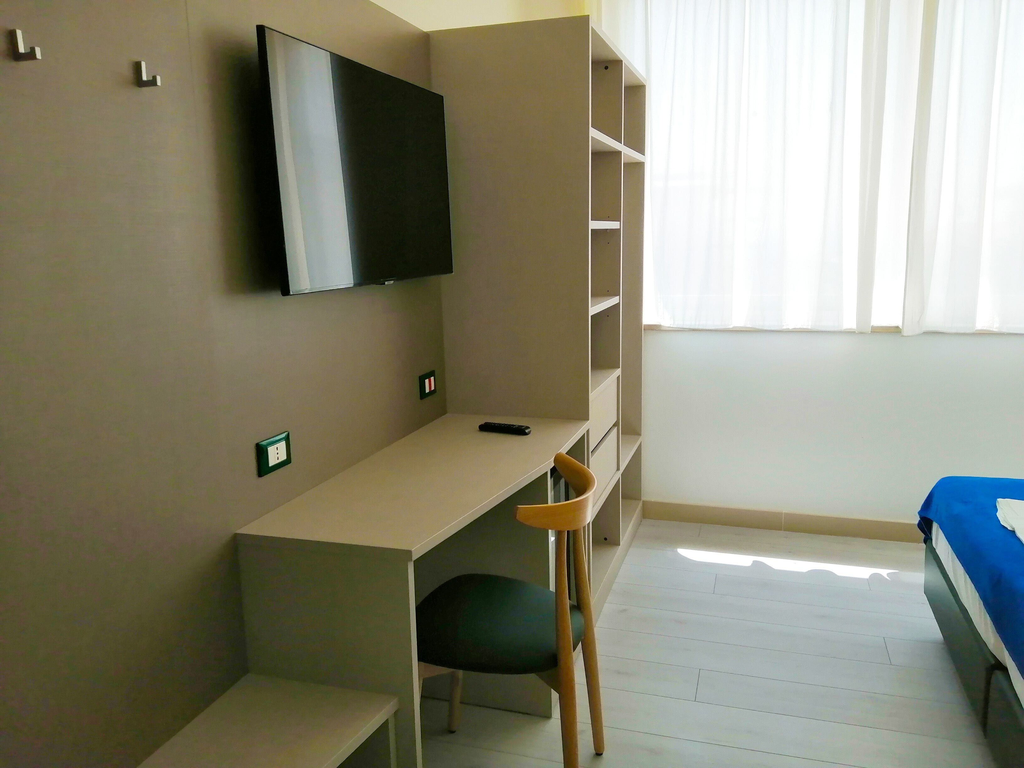 A bedroom with a bed, a desk, a chair, a TV mounted on the wall, and a window with curtains.