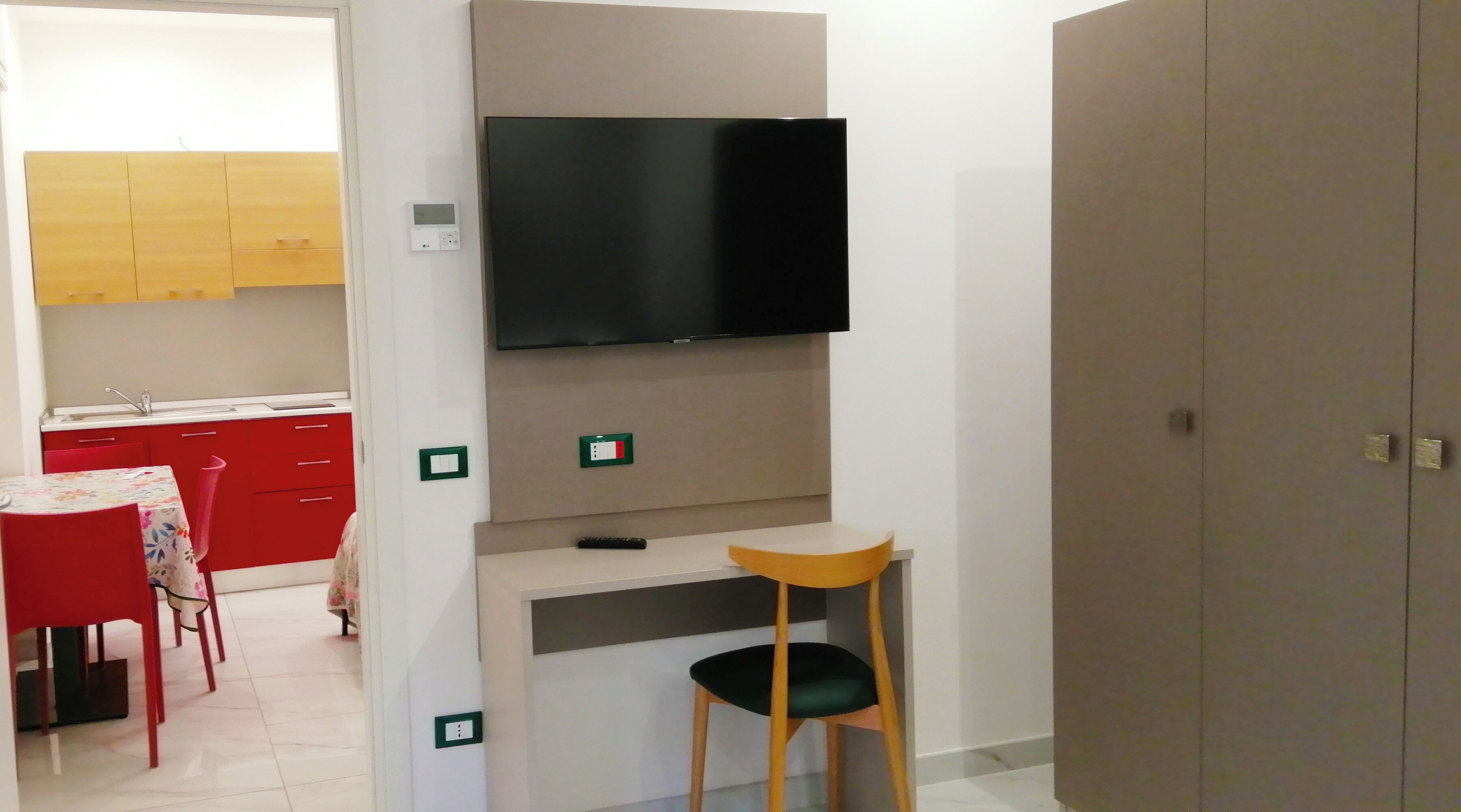 A compact room with a kitchenette, a TV mounted on the wall, and a small desk with a chair.