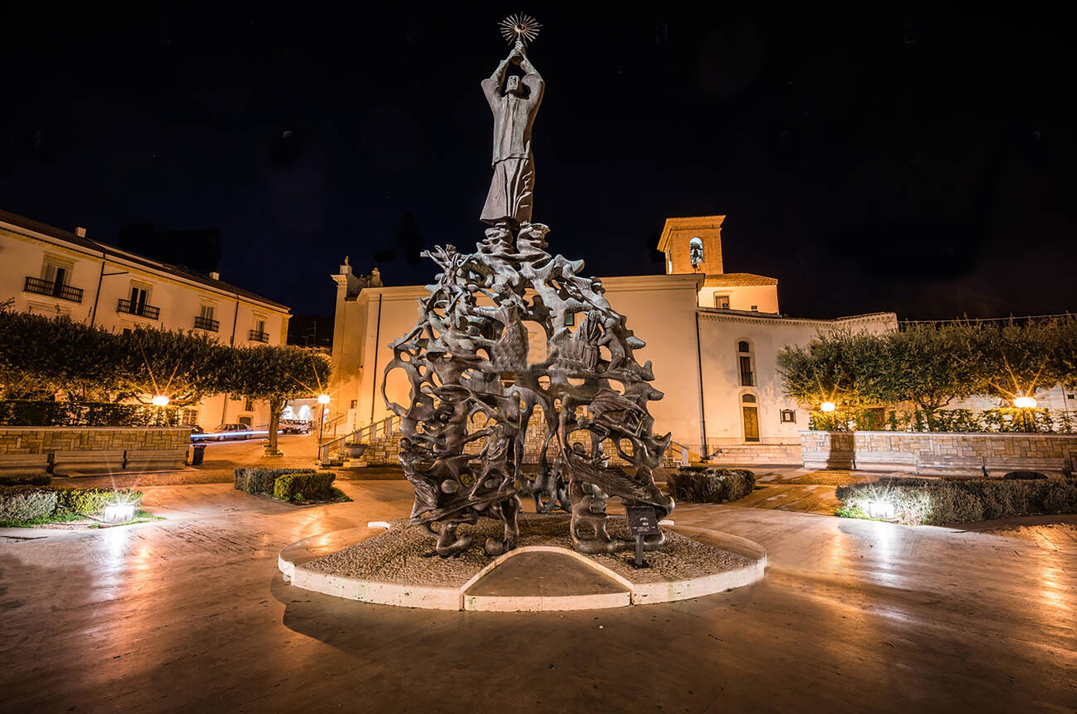 A nighttime outdoor sculpture in a plaza with surrounding buildings.