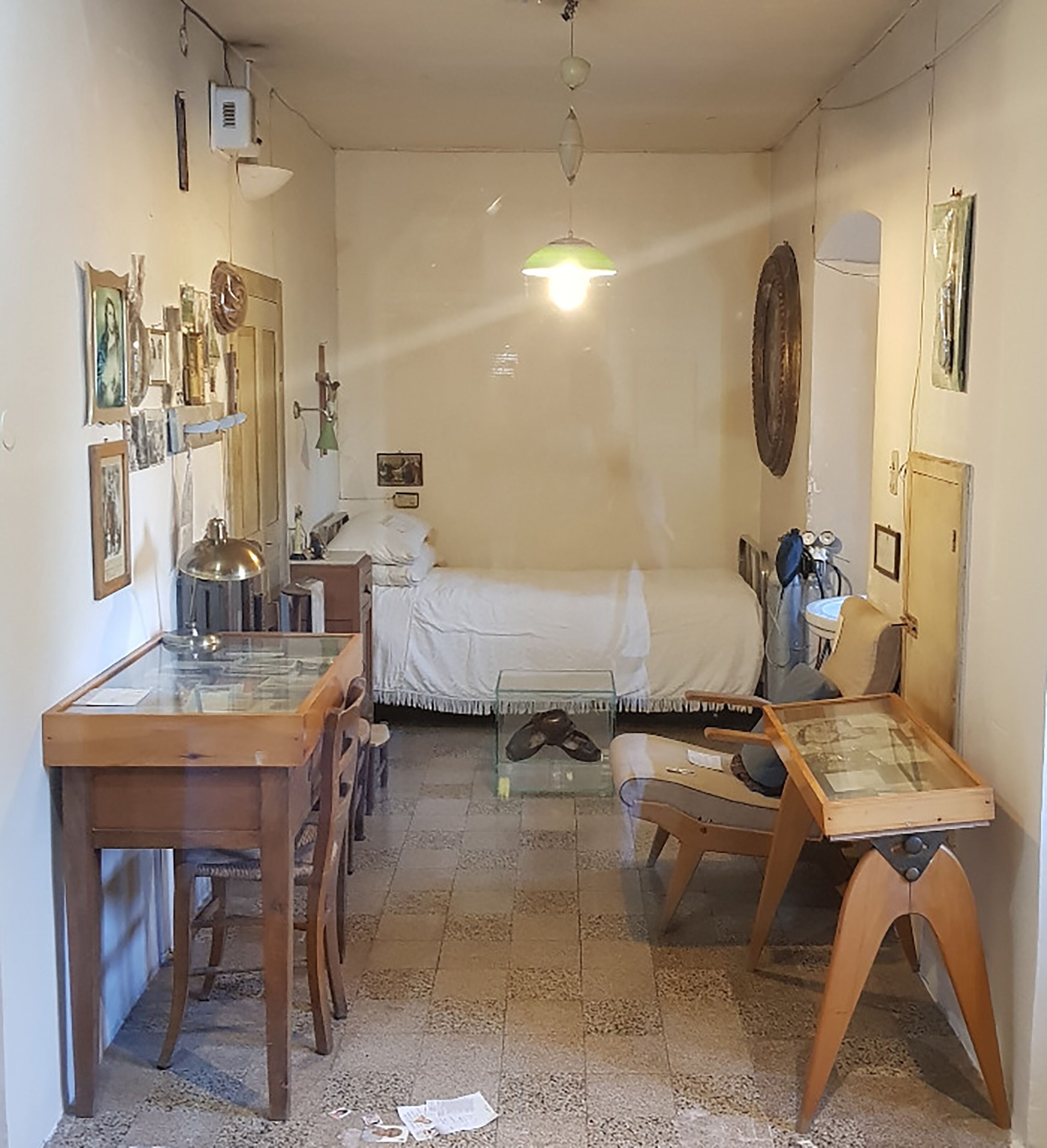A small, narrow room with a bed, a desk, and a chair. There are various items on the walls and a ceiling fan.