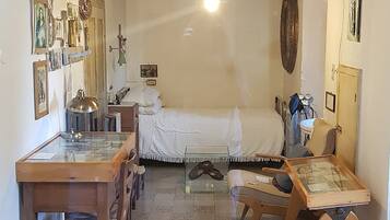 A small, narrow room with a bed, a desk, and a chair. There are various items on the walls and a ceiling fan.