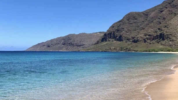 Ein Strand mit klarem Wasser, sandigem Ufer und hügeliger Kulisse im Hintergrund.