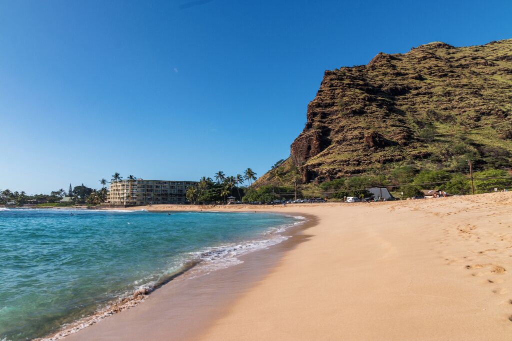 A beach with clear blue water, a sandy shore, and a building with multiple floors in the background.