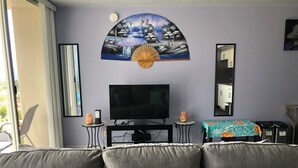 Ein Wohnzimmer mit einer Couch, einem Fernseher, einem kleinen Tisch mit Ventilator und einem Bild an der Wand.