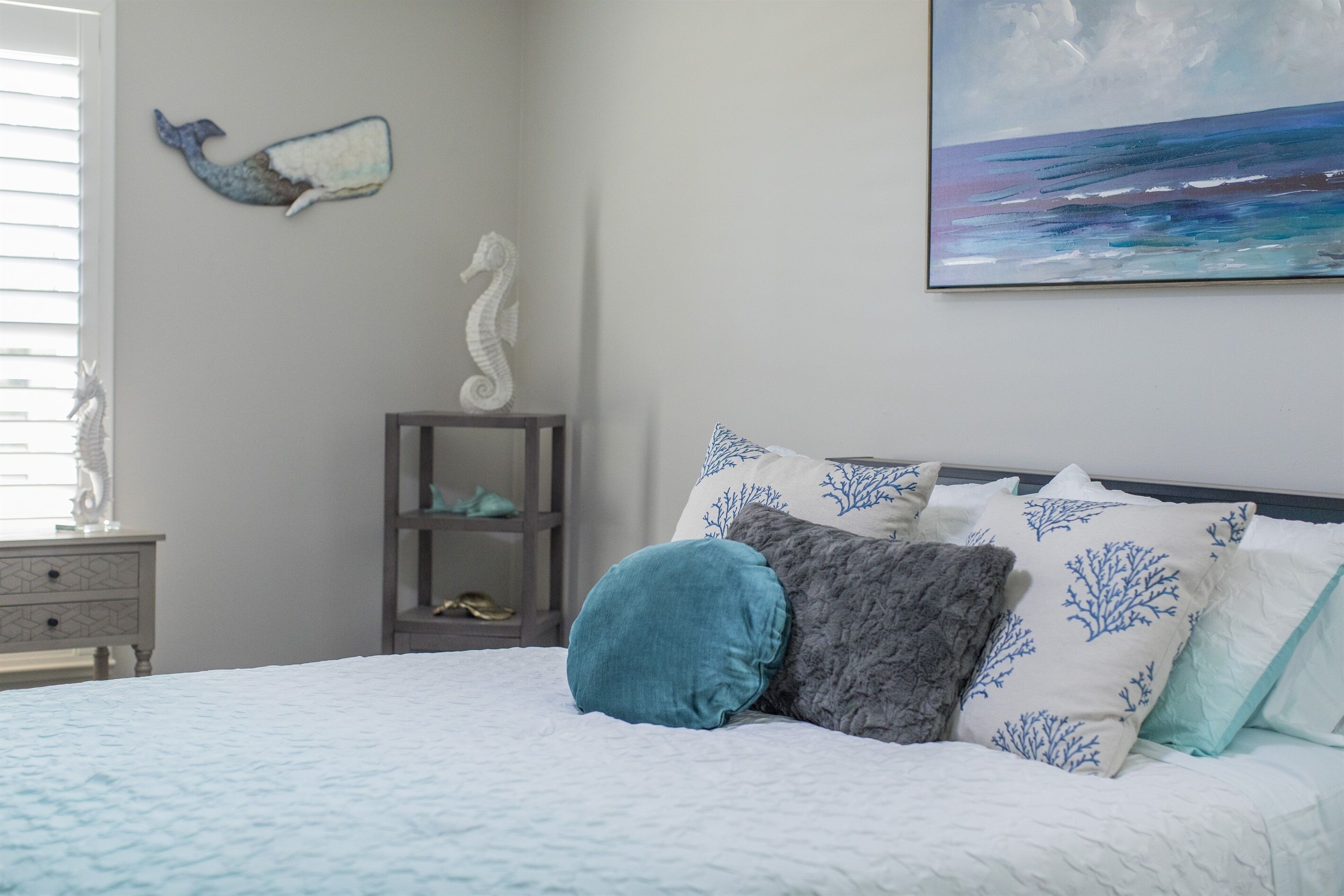 Une chambre à coucher avec un lit, des oreillers, une décoration représentant un hippocampe, une étagère avec des chaussures et un tableau représentant l’océan.