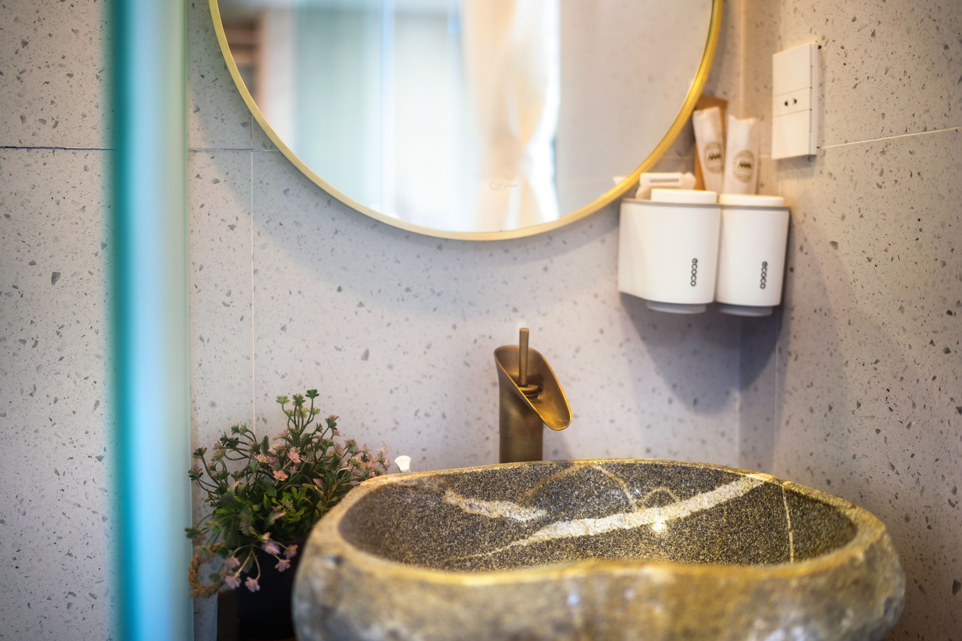 A bathroom with a round stone sink, a gold faucet, a mirror, and a wall-mounted soap dispenser.