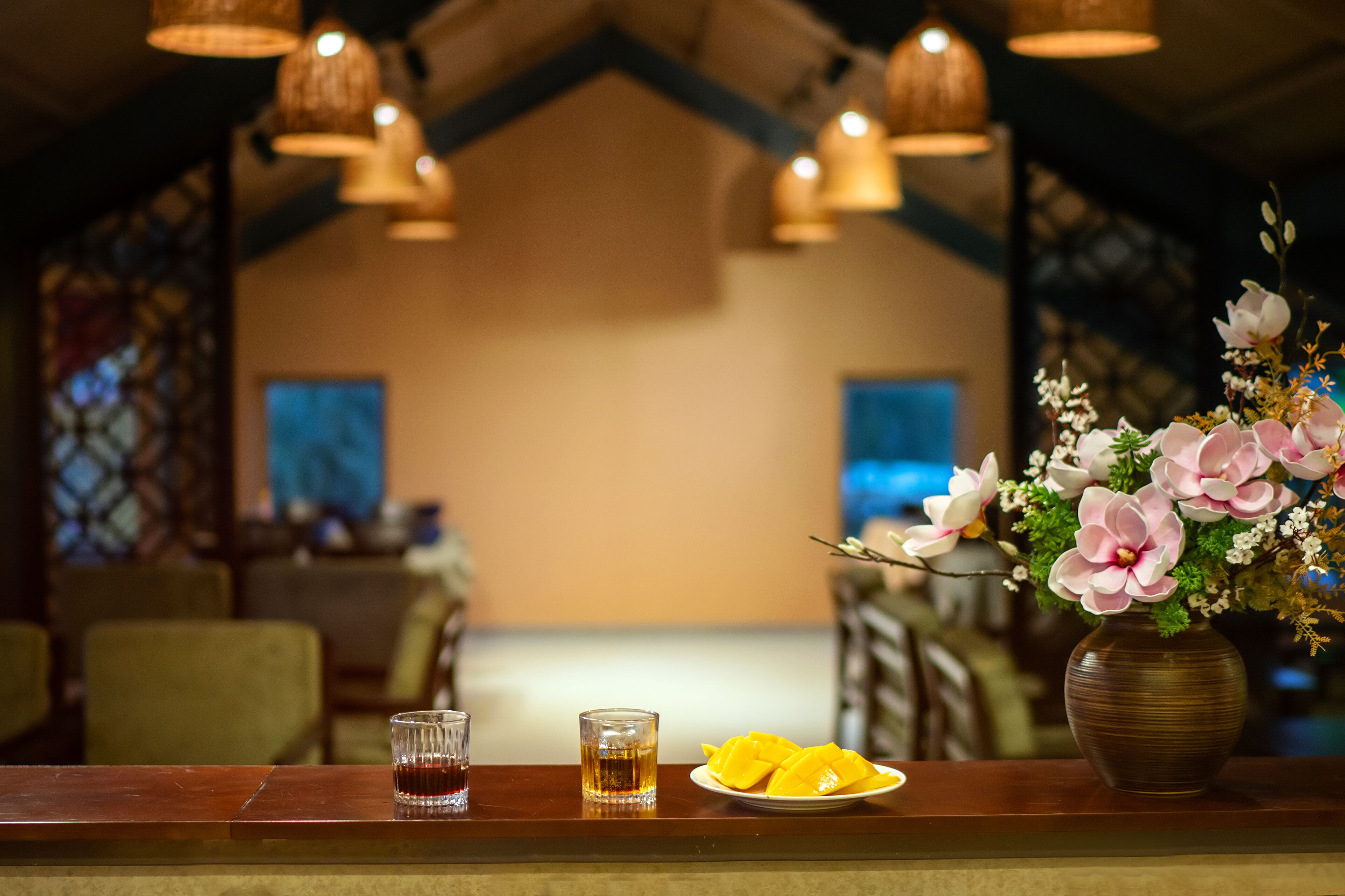 A restaurant interior with a wooden counter, two glasses of amber liquid, a plate of yellow fruit, and a vase with pink flowers.