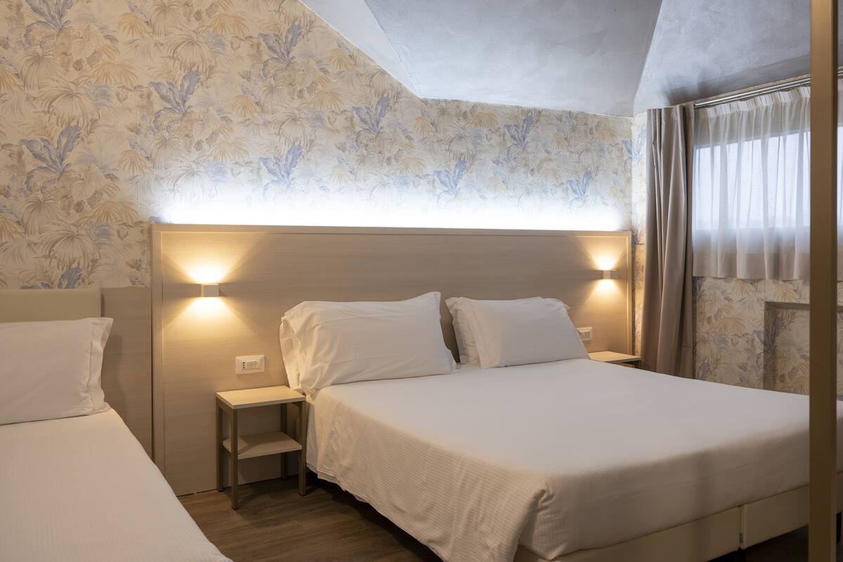 A hotel room with two beds, a nightstand, and a headboard with integrated lighting.
