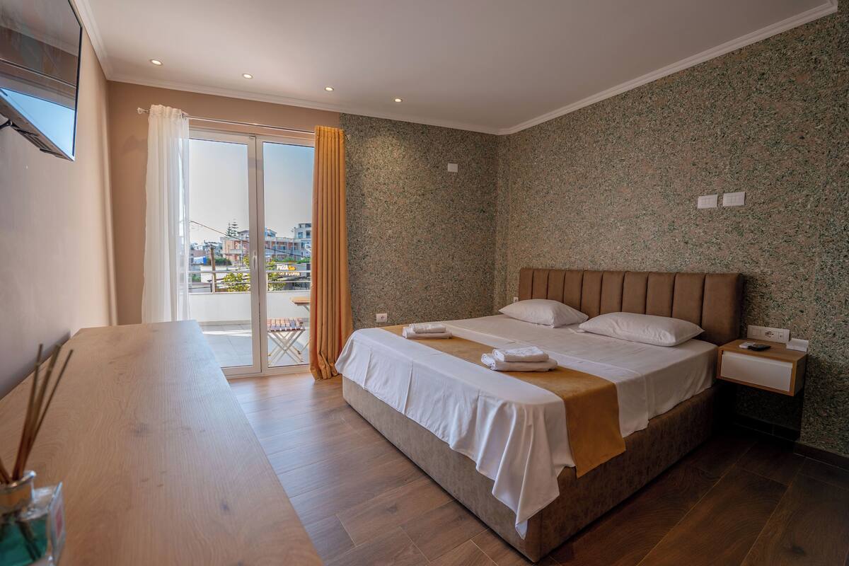 A modern hotel room with a large bed, a wooden desk, and a balcony with curtains.