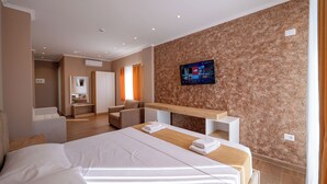 Ein modernes Hotelzimmer mit einem großen Bett, einem Holztischchen, einem flachbildfernseher an der Wand und einer Sitzecke mit Sofa und Sesseln.