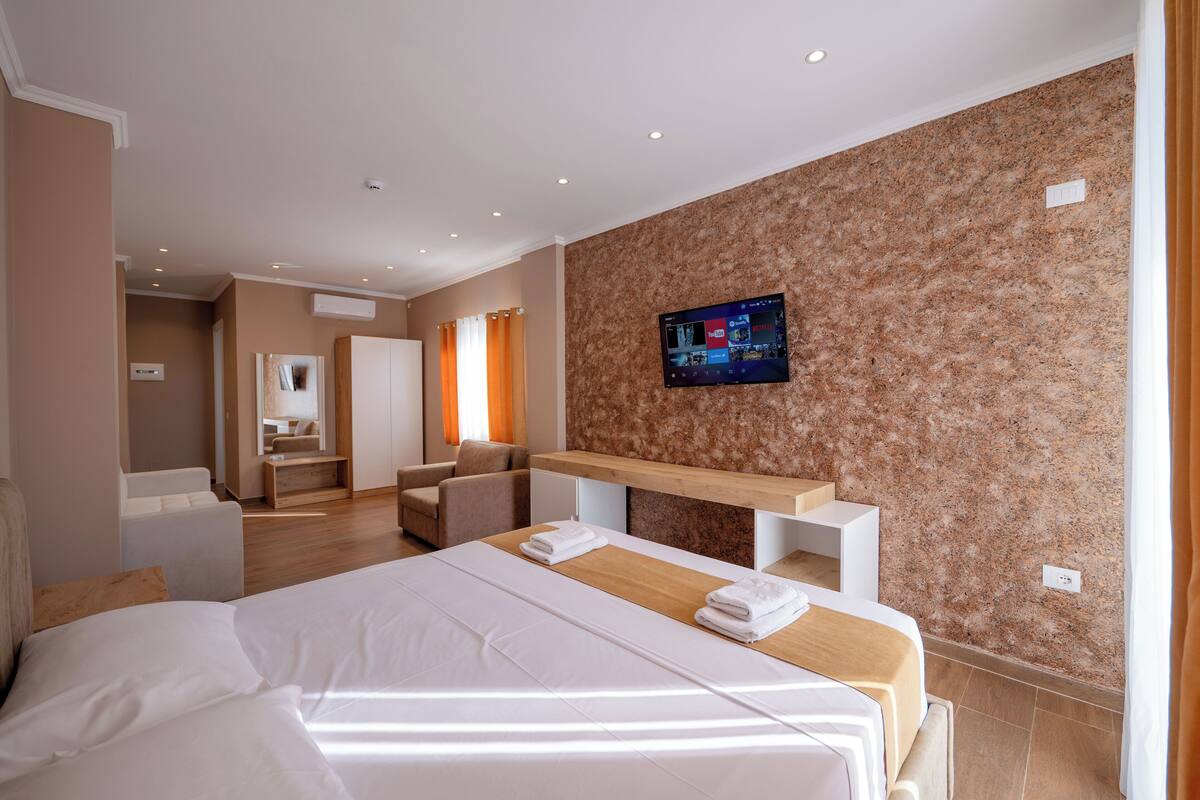 A modern hotel room with a large bed, a wooden side table, a flat-screen TV mounted on the wall, and a seating area with a sofa and armchair.