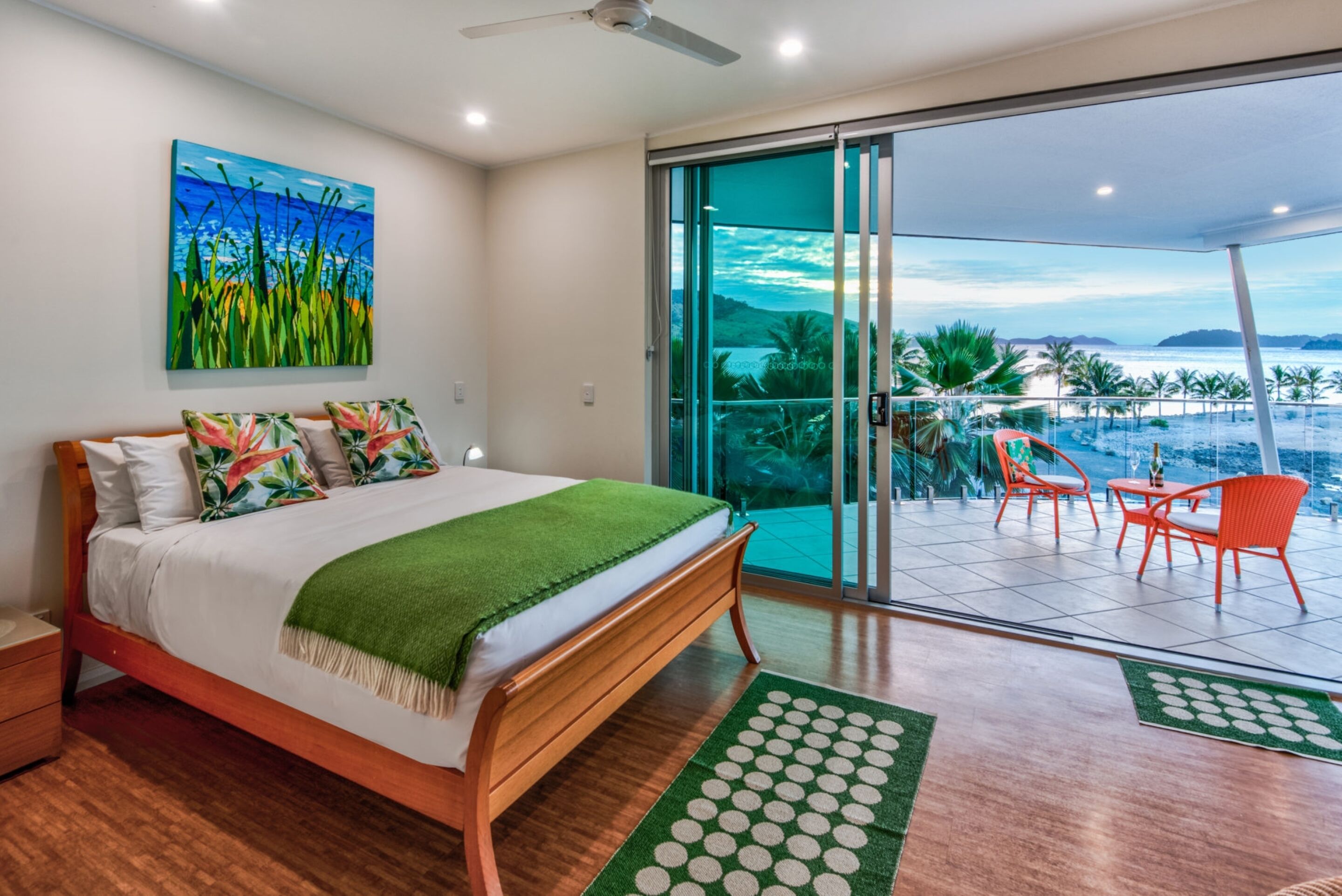 A bedroom with a bed, a painting, a balcony with chairs, and a view of the ocean.