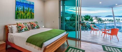 A bedroom with a bed, a painting, a balcony with chairs, and a view of the ocean.