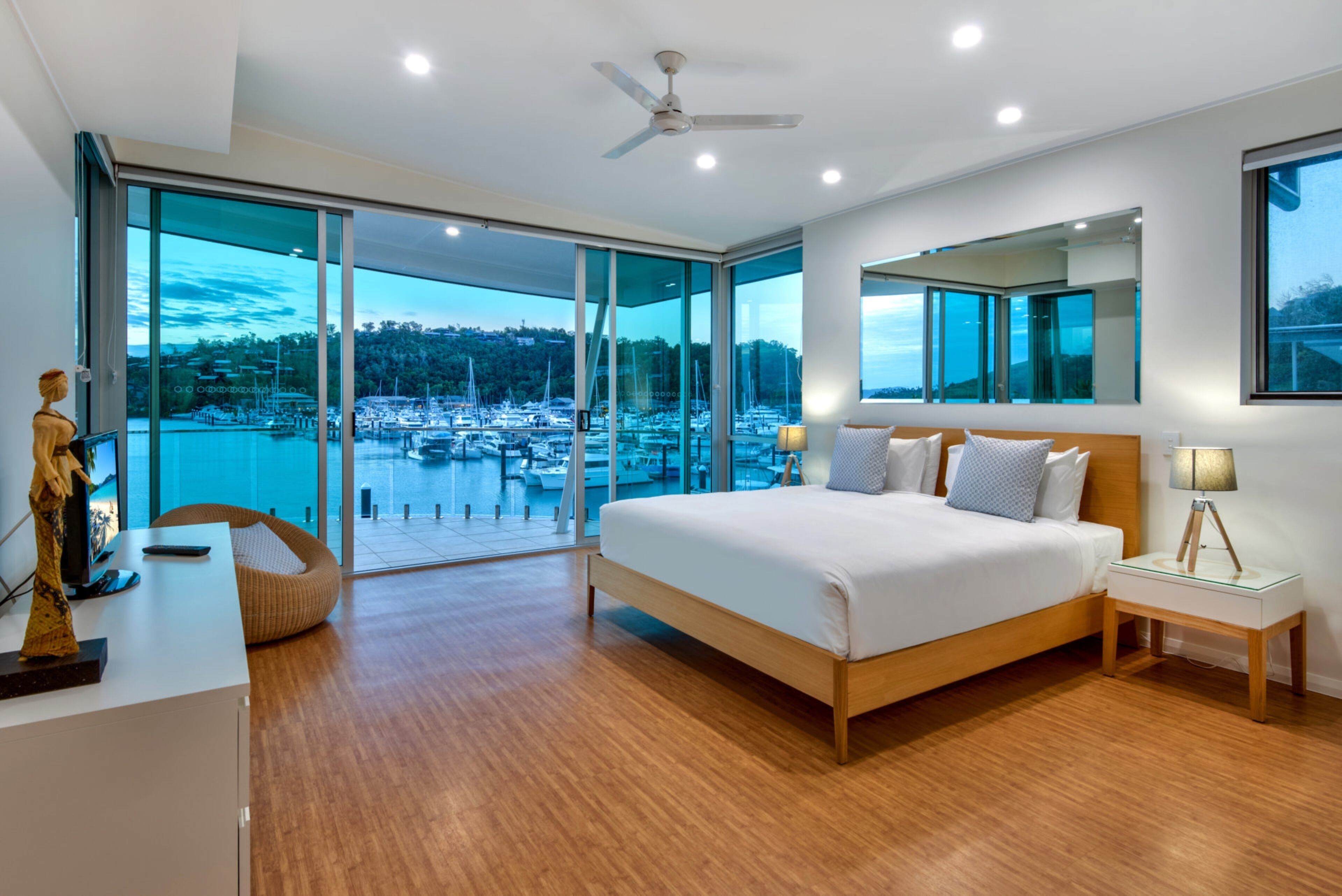 A modern bedroom with a large bed, a desk, and a view of a marina through floor-to-ceiling glass doors.