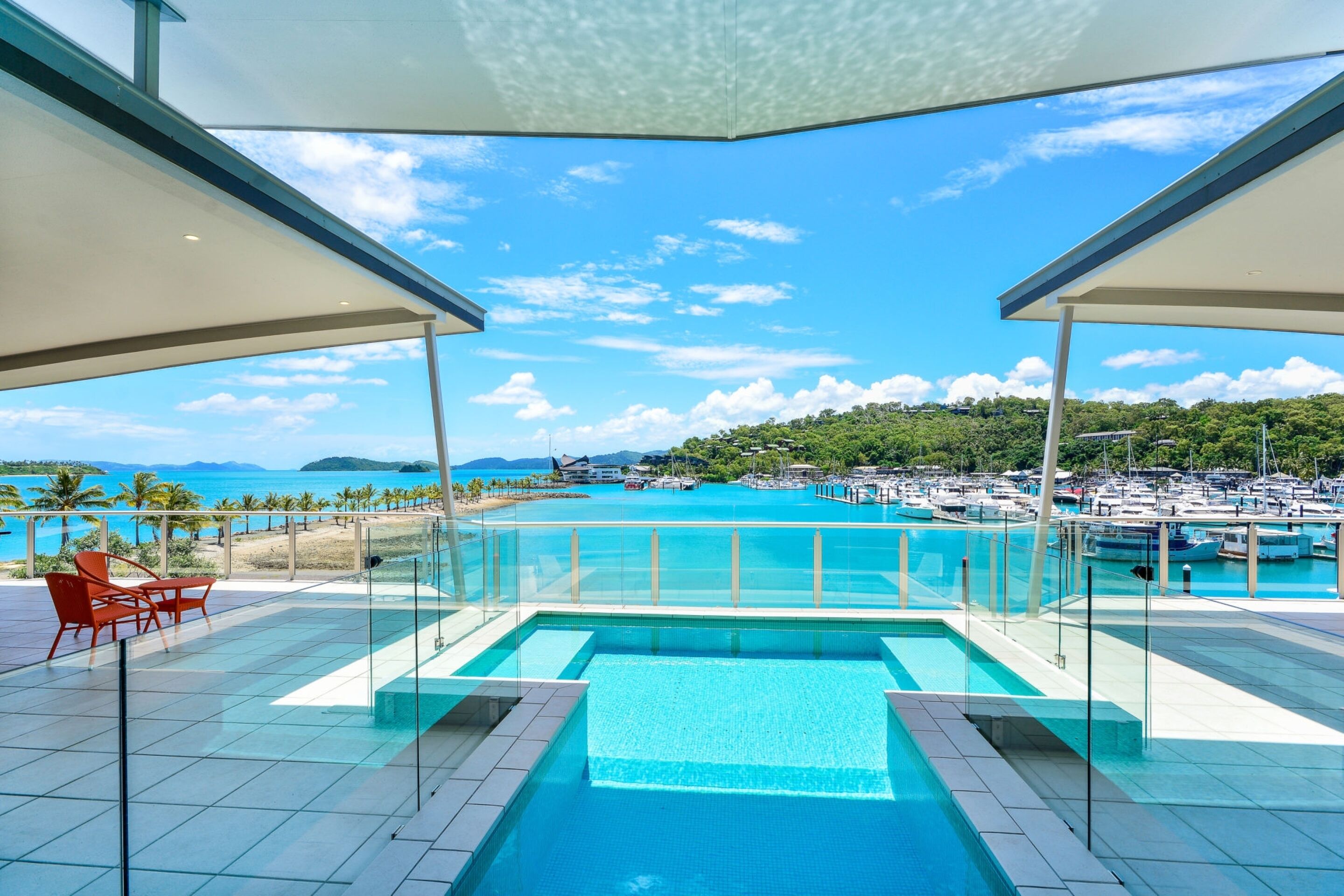 A swimming pool with clear blue water, surrounded by a glass railing, and overlooking a marina with boats and a distant coastline.