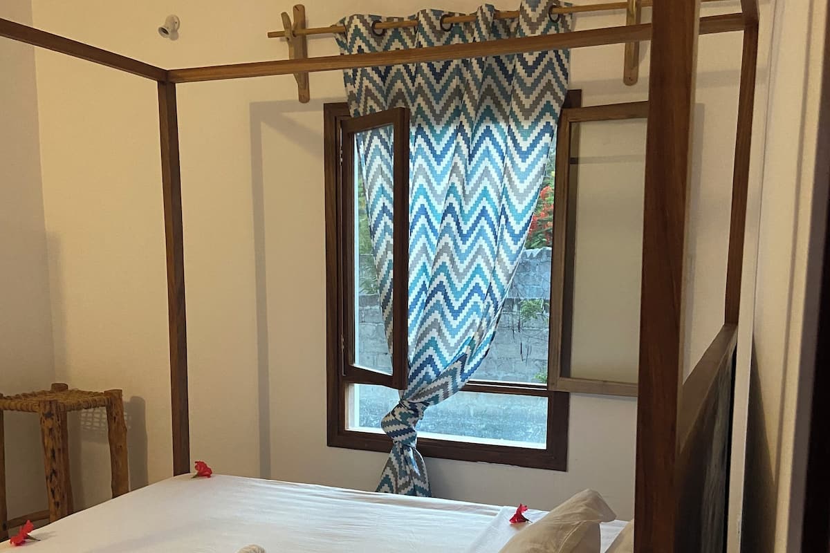 Una cama bien tendida con sábanas blancas y flores rojas, un cabecero de madera y una ventana con cortina de estampado en zigzag.
