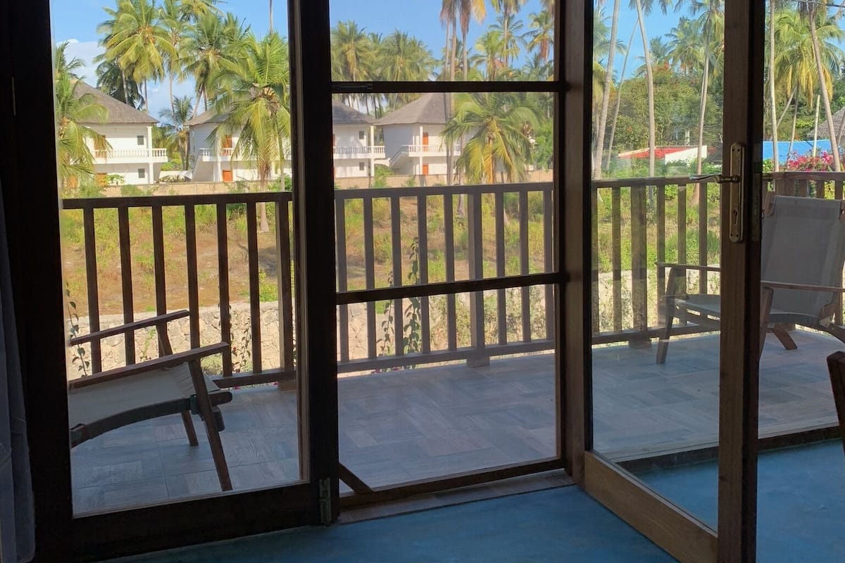 Una habitación con un gran ventanal corredera que da a un balcón con vistas a palmeras y casas.