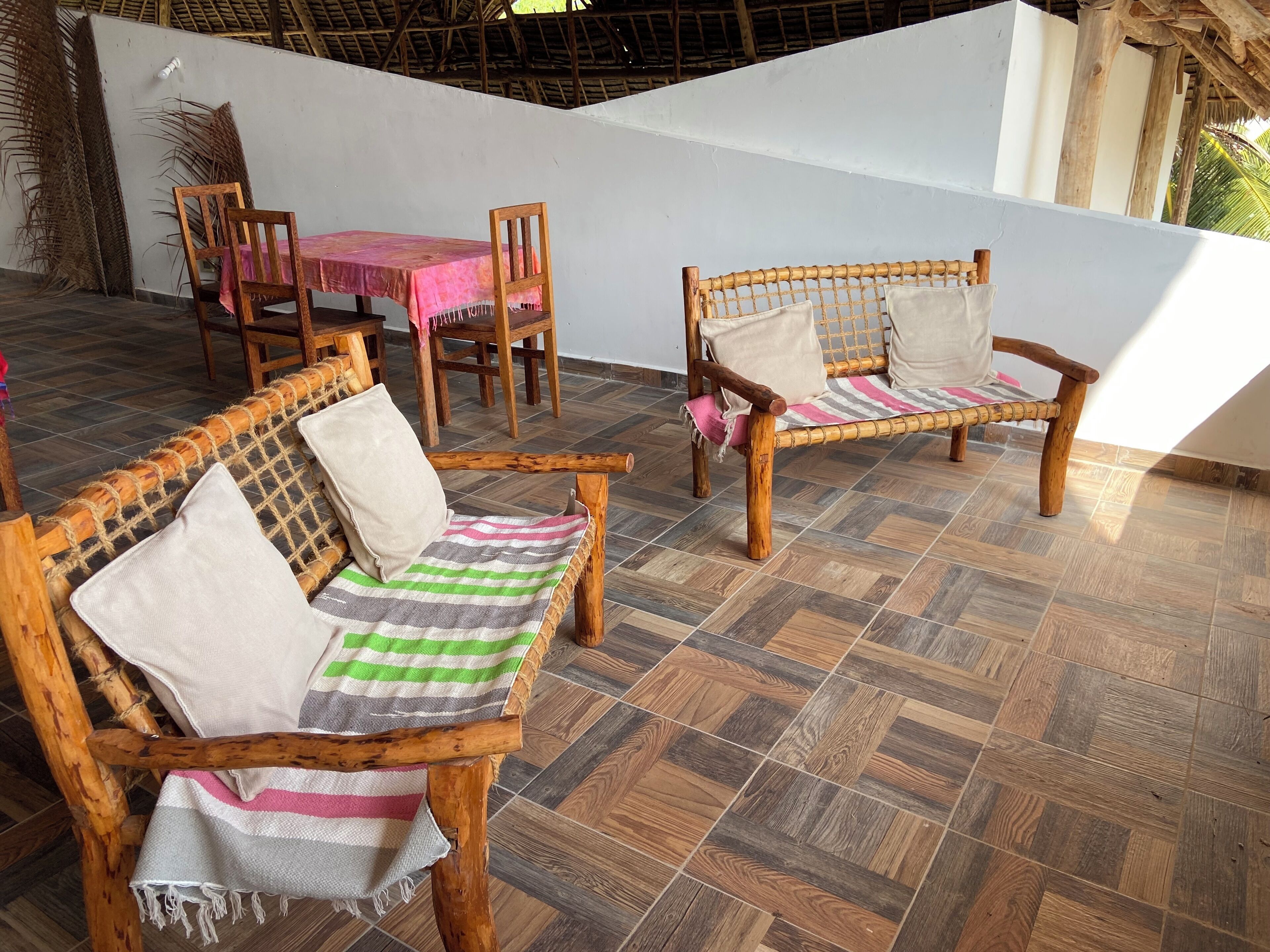 Uma área de pátio com mobiliário de madeira, incluindo dois sofás de vime e uma mesa com cadeiras, sobre um piso de azulejos estampados.