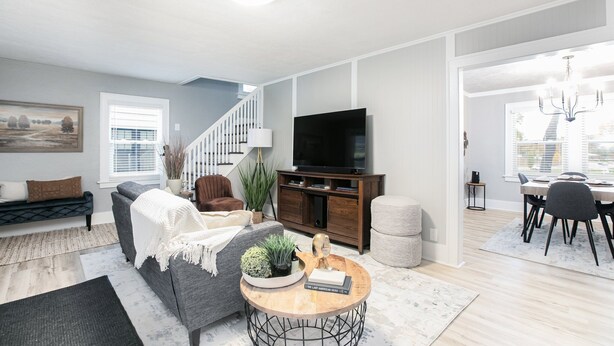 Una sala de estar moderna con un sofá gris, una mesa redonda de centro con objetos decorativos, un televisor sobre un mueble de madera y una escalera que lleva a otro nivel.