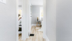 Un pasillo estrecho con paredes blancas y suelo de madera, que conduce a un dormitorio con cama, mesita de noche y ventana.