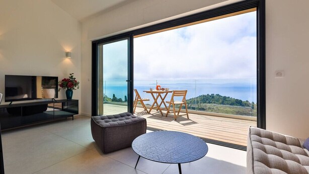A modern living room with a TV, sofa, and a table with chairs, overlooking a scenic view through large glass doors. - Large 2-room apartment with panoramic sea view (Rayol-Canadel-sur-Mer)
