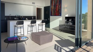 A modern kitchen with a bar area, a dining space with chairs, a sofa, and a large window. - Large 2-room apartment with panoramic sea view (Rayol-Canadel-sur-Mer)