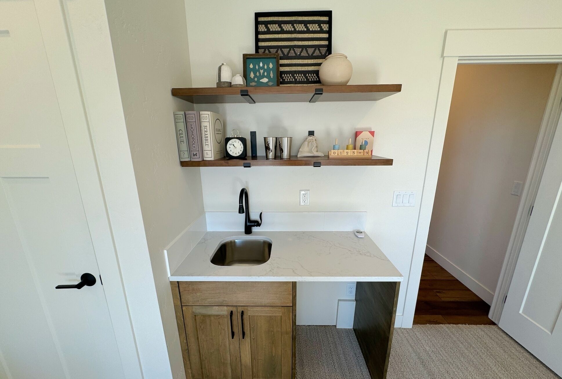 A compact kitchen with a sink, wooden cabinets, and two shelves above containing books, a clock, and decorative items.