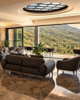 Una sala de estar moderna con un sofá grande, sillones y una mesa de centro. Un ventanal ofrece vistas a una montaña boscosa.