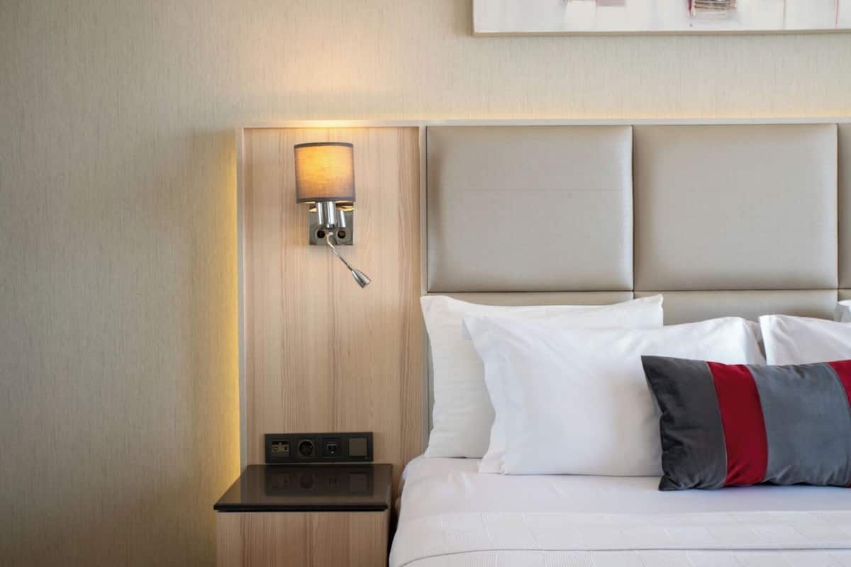 A hotel room with a bed, white bedding, a nightstand with a lamp, and a wall-mounted control panel.