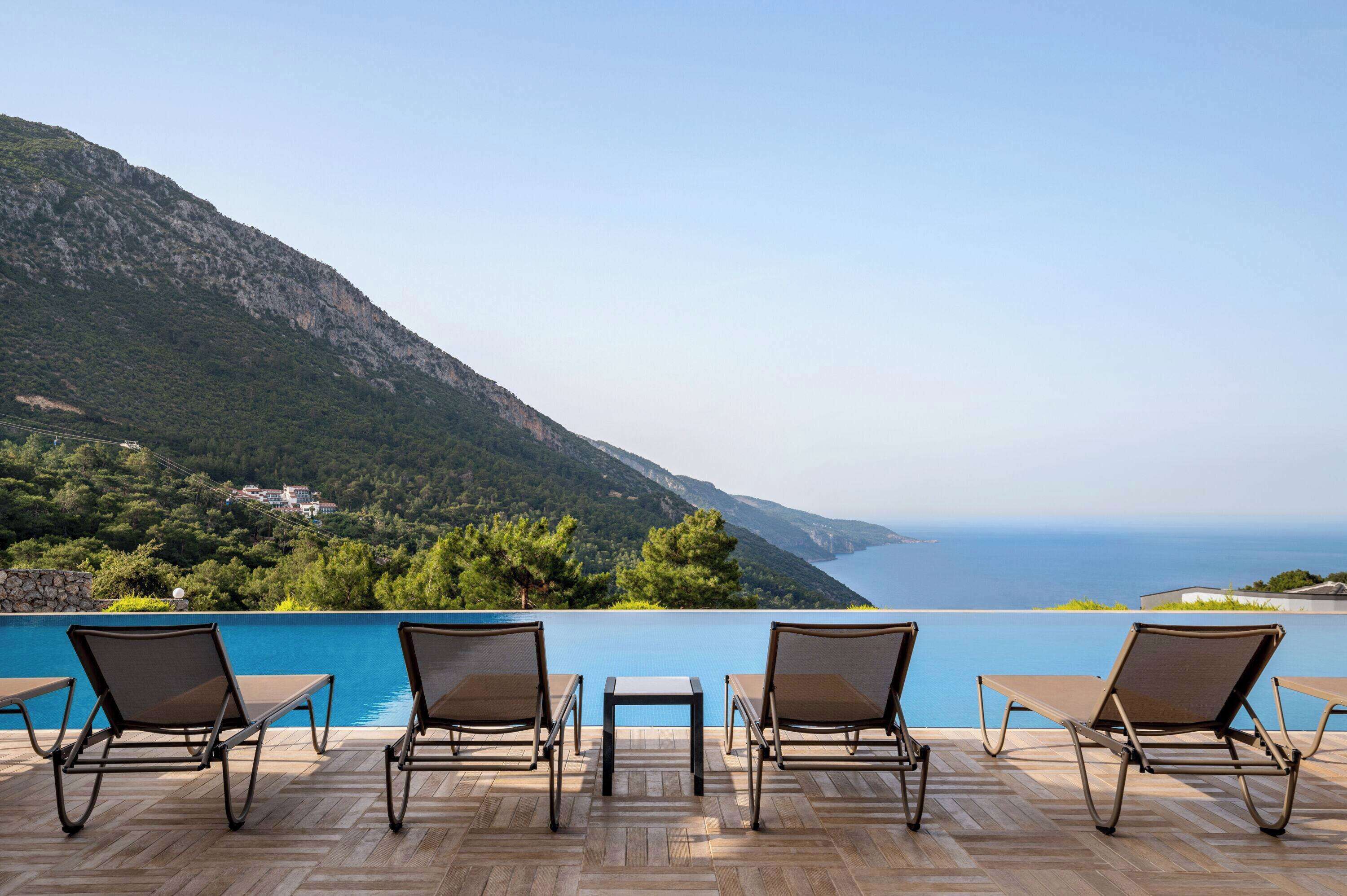 A poolside area with lounge chairs and a table overlooking a mountainous landscape and the sea.
