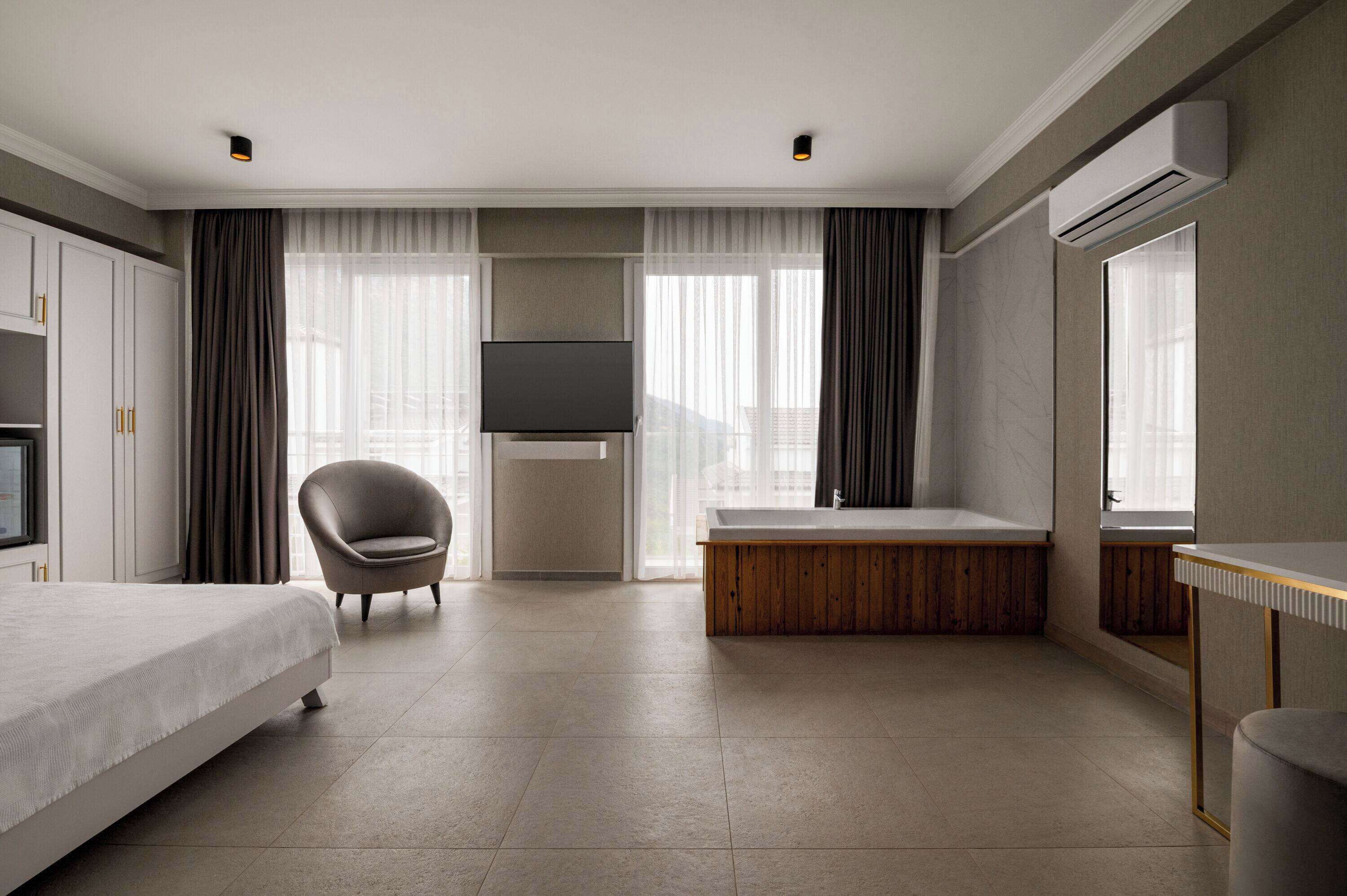 A modern hotel room with a bed, a chair, a large window with curtains, a flat-screen TV, a wooden bench, and an air conditioning unit.