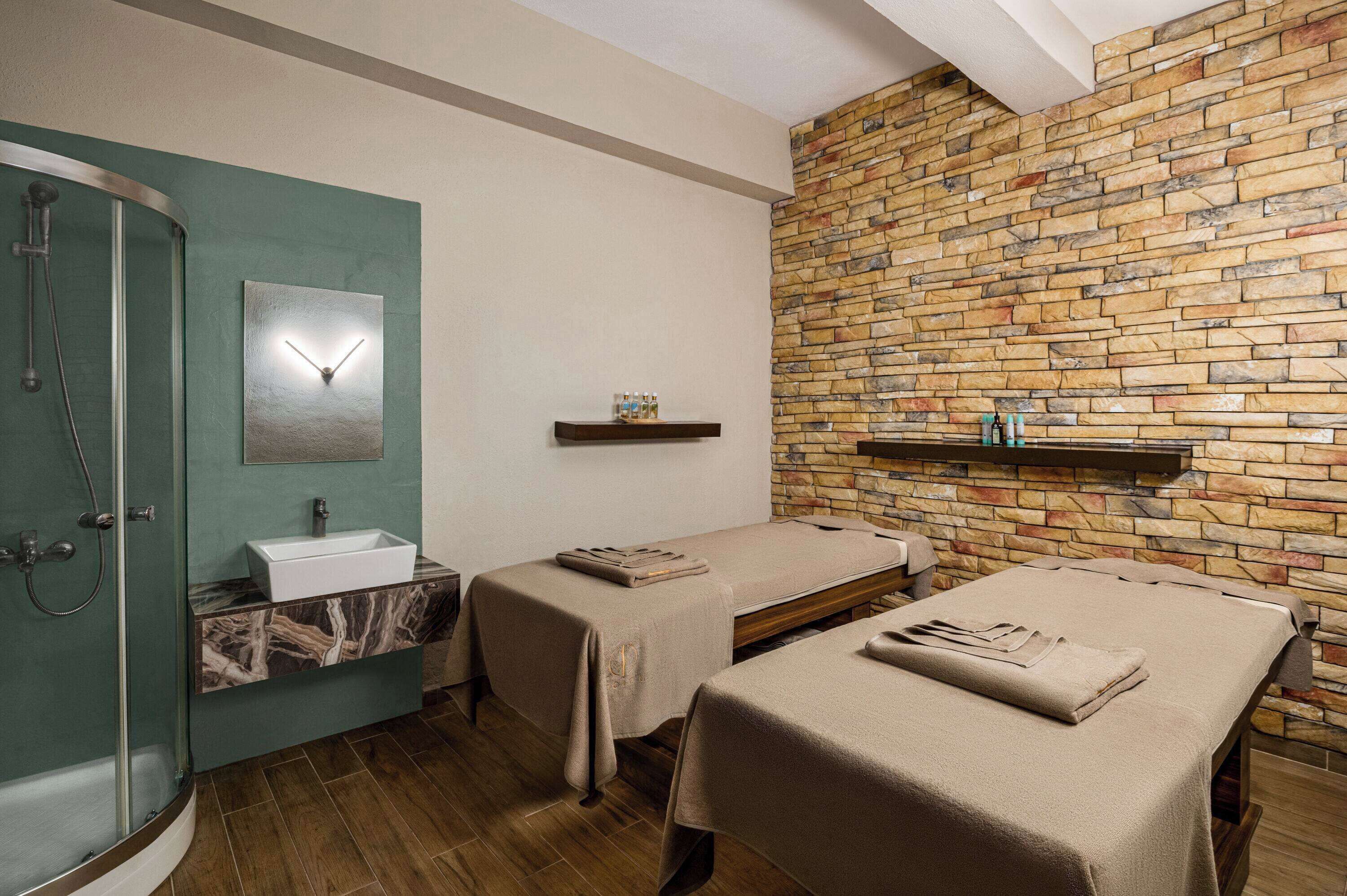 A spa room with a stone wall, a mirror, a sink, and two massage tables.