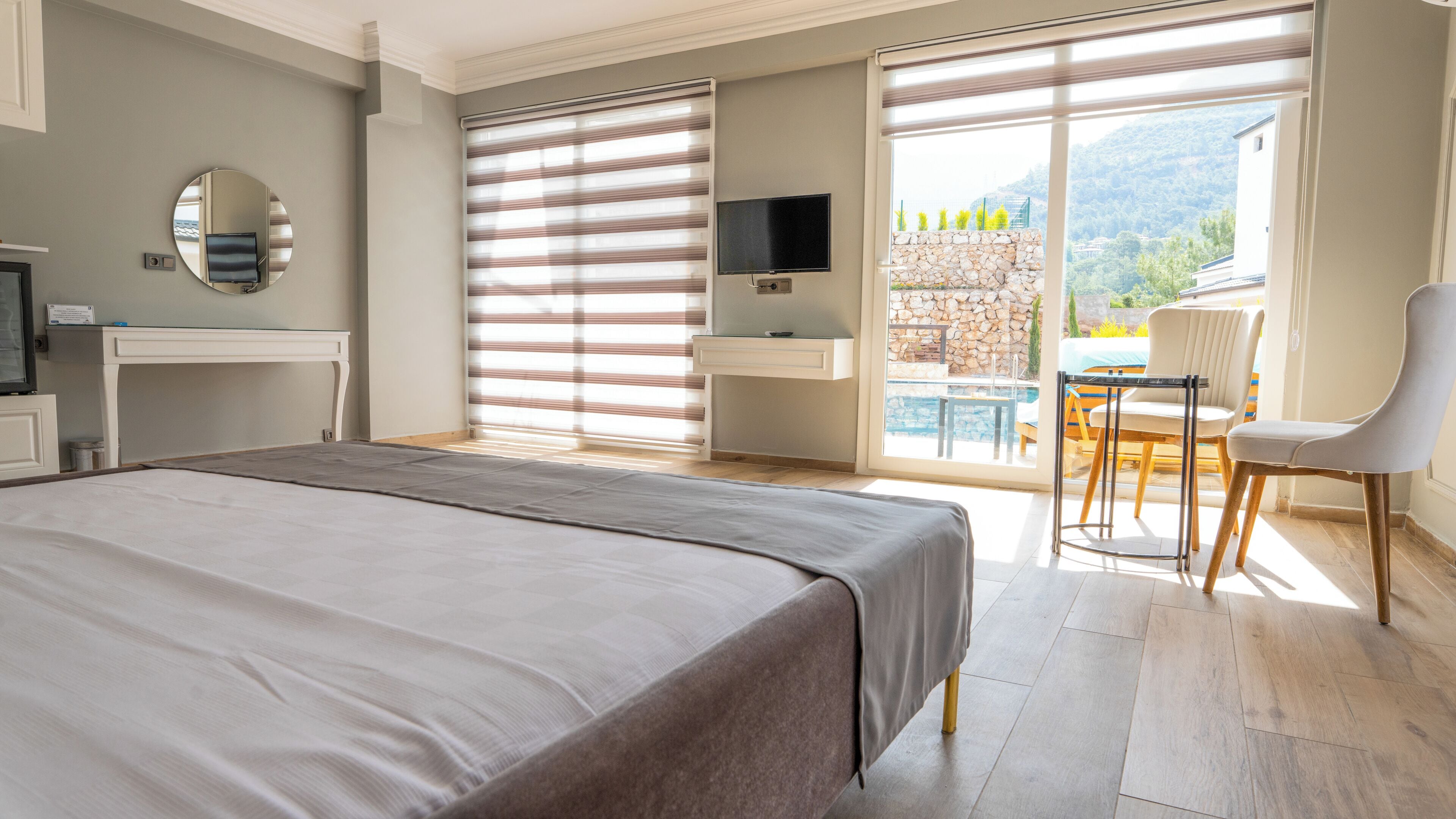 A modern hotel room with a large bed, a television, a mirror, a bathroom with a stone wall, and a balcony with a pool and chairs.