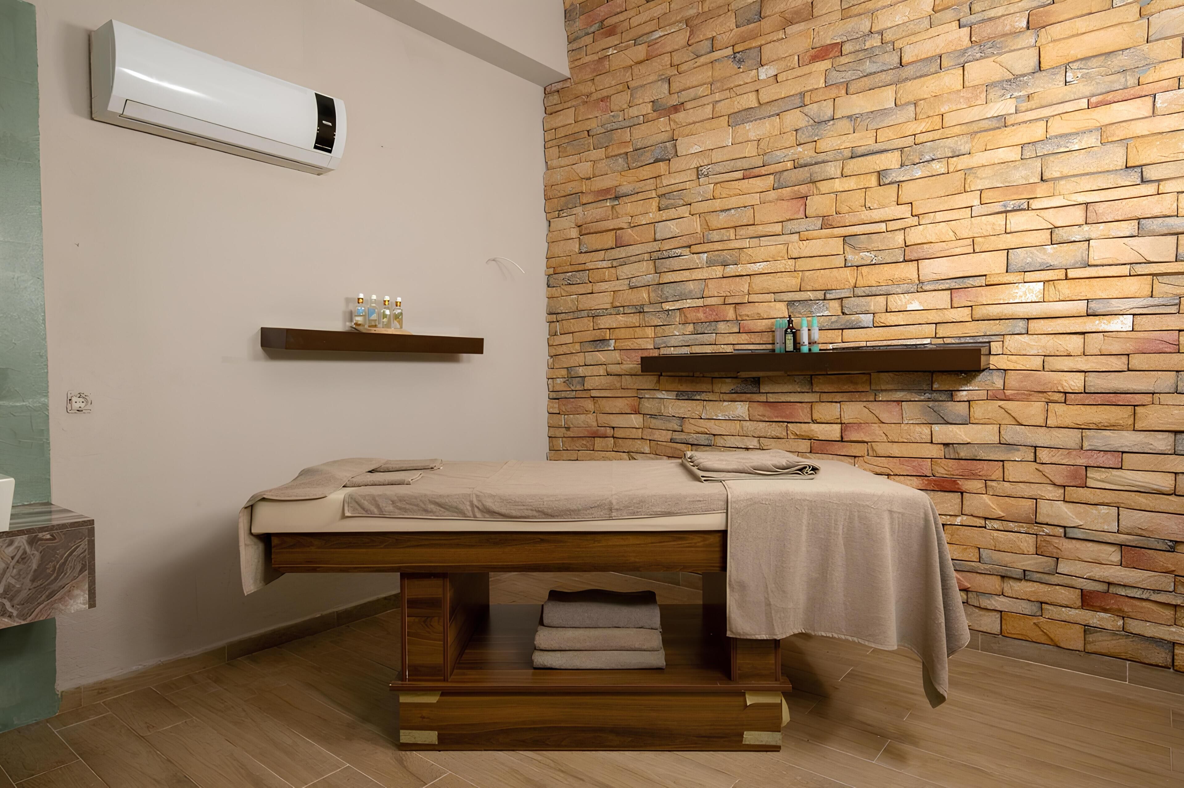 A room with a massage table, stone wall, wooden floor, and a shelf with bottles.