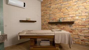 Una habitación con camilla de masaje, pared de piedra, suelo de madera y estante con botellas.