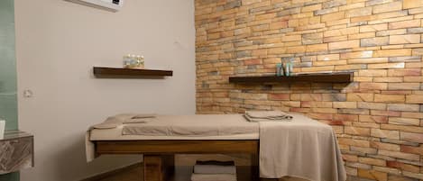A room with a massage table, stone wall, wooden floor, and a shelf with bottles.