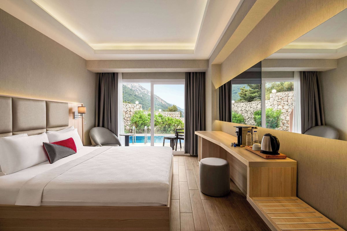 A hotel room with a large bed, a desk, a chair, a small stool, and a view of a pool and mountains.