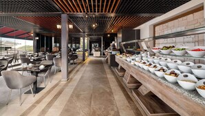 A modern dining area with a long counter displaying various food items, a ceiling with a grid pattern, and a view of the outdoors. - Ramada by Wyndham Fethiye Oludeniz (Fethiye)