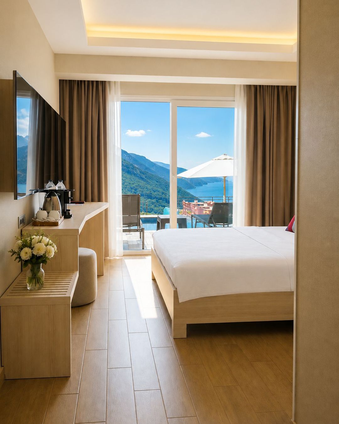 Habitación de hotel con cama, escritorio, silla, una mesita, un jarrón de flores, un ventanal con vistas a montañas y un lago, y una puerta corrediza que da a un espacio exterior.