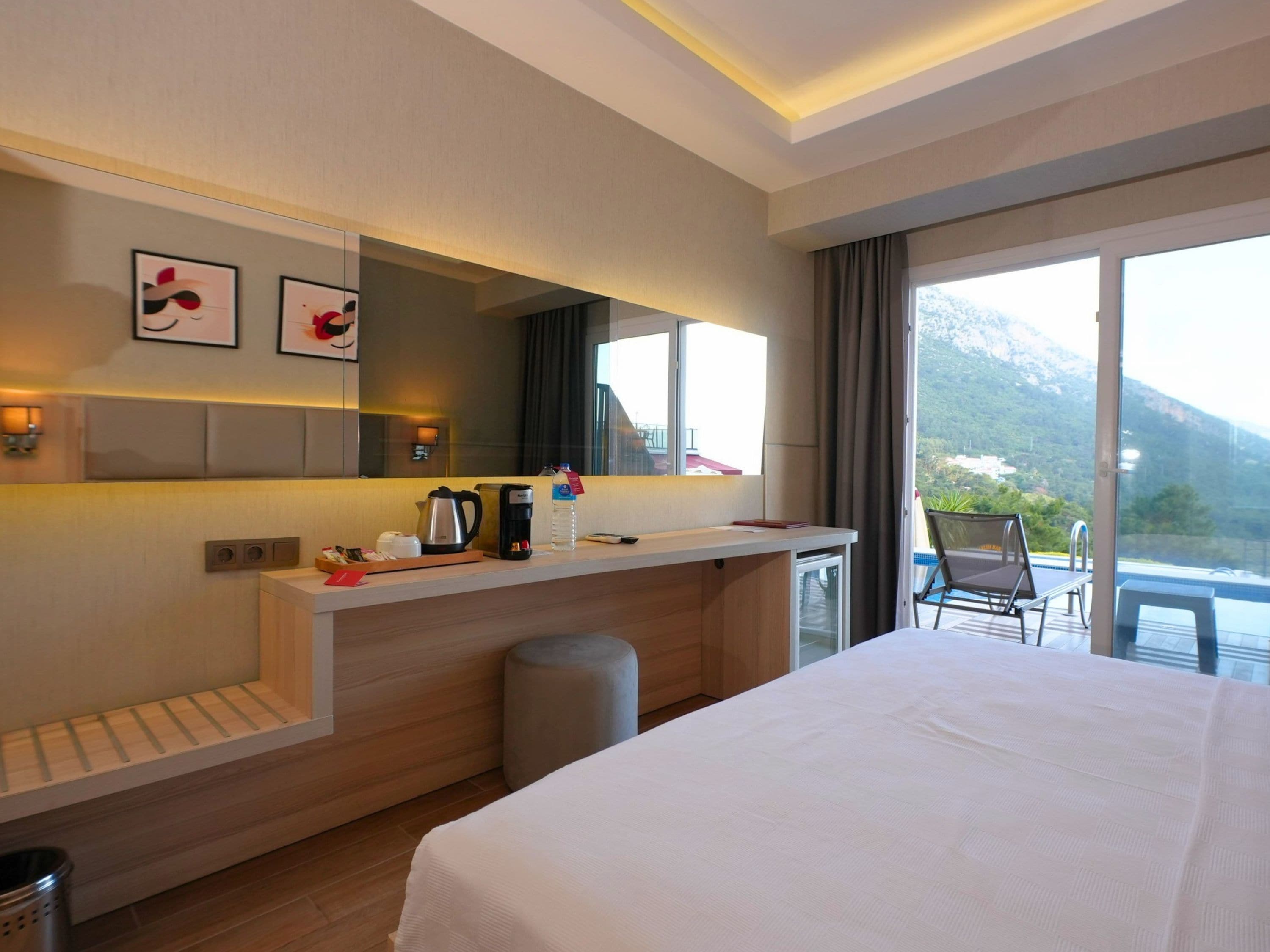 Habitación de hotel con cama, escritorio con cafetera, una silla, un taburete pequeño y vistas a una montaña a través de un ventanal grande.